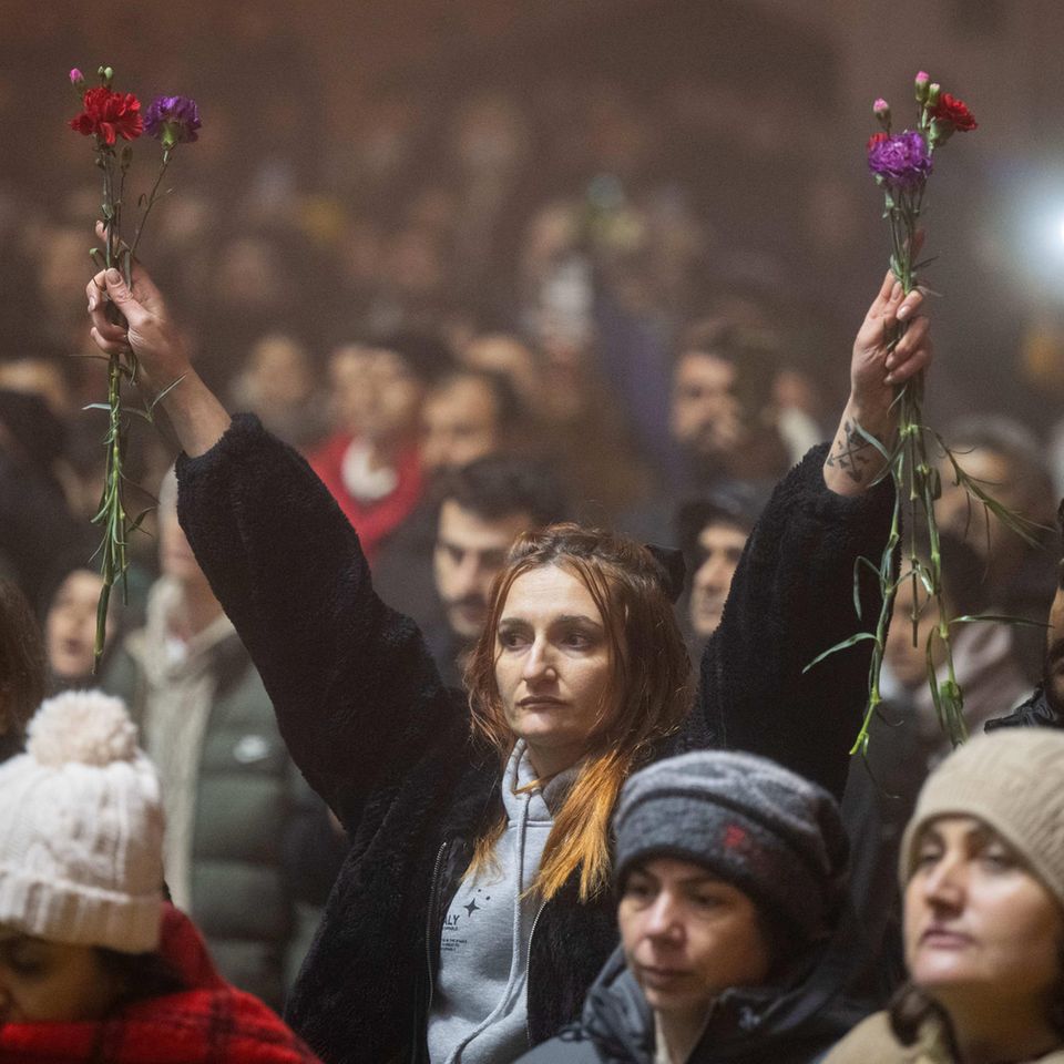 Antakya, Türkei. Inmitten tausender Menschen hält eine Frau bei einer Kundgebung am Jahrestag des Erdbebens Blumen in die Höhe. Vor einem Jahr hatten mehrere zehntausend Menschen bei einem massiven Erdbeben im Südosten der Türkei ihr Leben verloren.